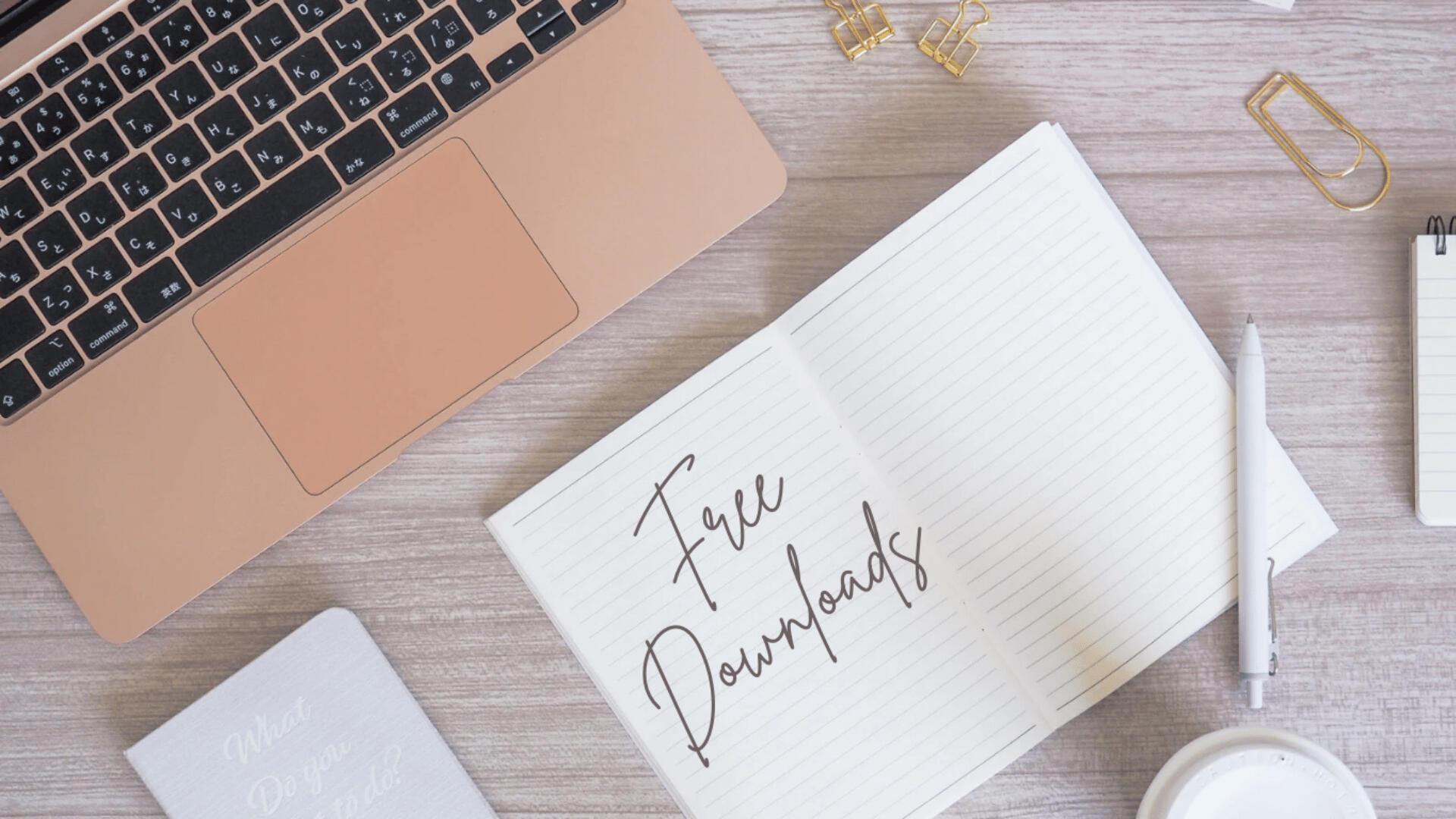 Free Writing Resources — Downloadable Tools for Aspiring Authors Flat-lay of a rose gold laptop, a lined notebook labeled “Free Downloads,” gold paper clips, a pen, and a soft neutral background. Represents access to digital writing tools and downloadable resources for aspiring authors and creatives.